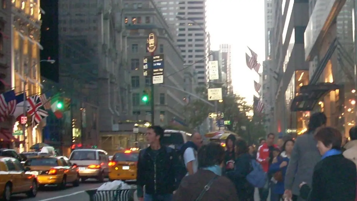 ニューヨークの五番街の昼間の街並み。ビルから複数のアメリカ国旗、イギリス国旗、ブラジル国旗などが掲揚されており、通りの両側には多くの人々が歩き、イエローキャブや他の車が行き交っています。