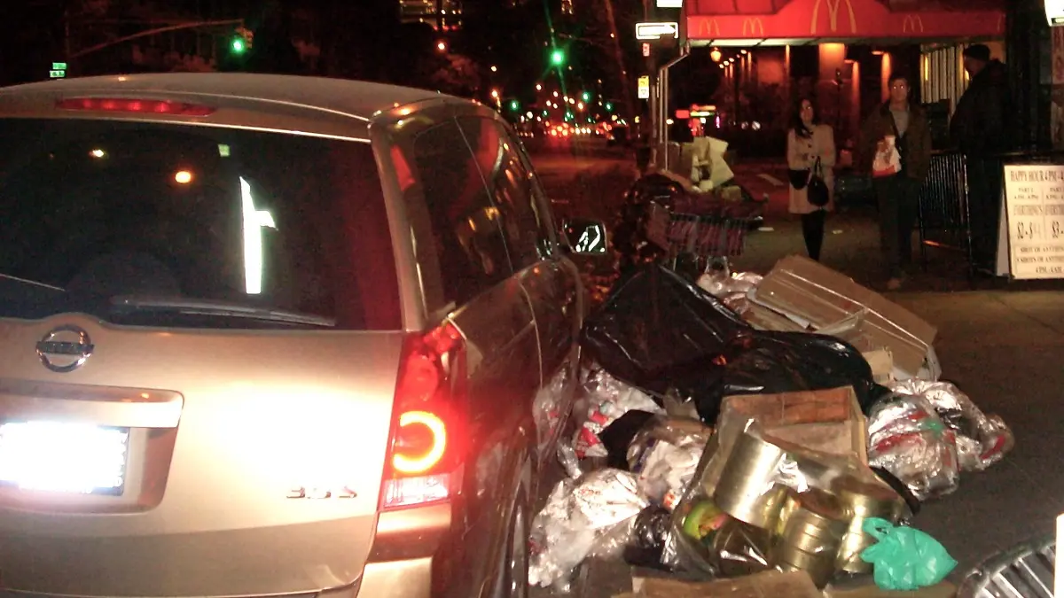 夜のニューヨークの街角。ごみ袋や段ボールが積み上げられた路肩に駐車されたシルバーのSUV車。背景には明るいネオンや街灯が瞬き、数人の通行人が行き交う、都会の喧騒と日常を捉えたアイキャッチ画像。