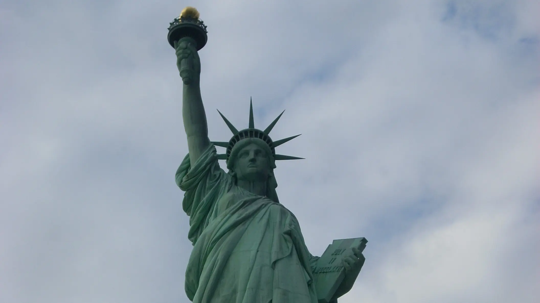自由の女神像をローアングルから撮影した迫力ある写真。女神はたいまつを掲げており、背景は曇り空。