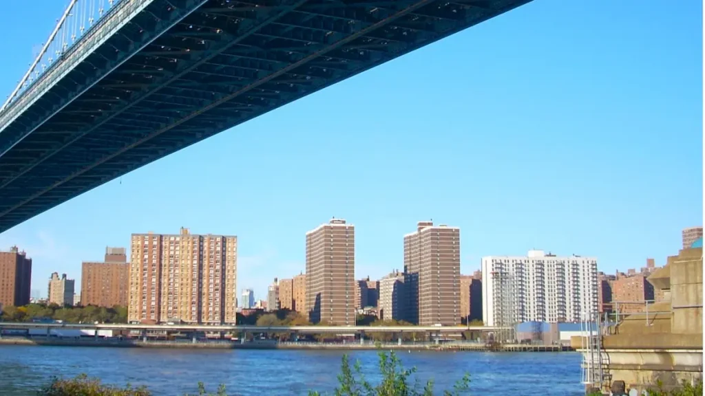 ニューヨークのマンハッタン橋の下からイースト・リバー越しに見る、ブルックリン(DUMBO地区)またはマンハッタン側の高層住宅群の眺め。橋の鉄骨構造が上部を覆い、川にはフェリーやタグボートのような船が浮かんでいる。