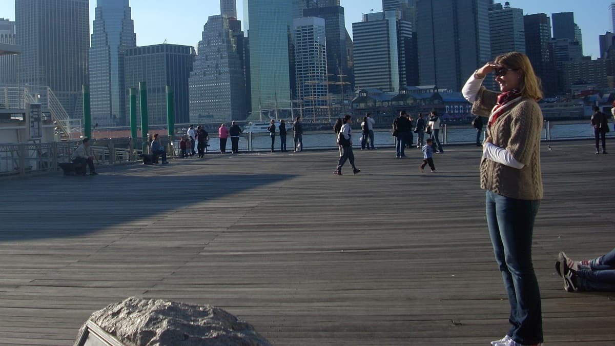 ニューヨークのサウスストリート・シーポートにある埠頭から撮影された、マンハッタンの高層ビル群の全景写真。人々がウッドデッキの上を歩いている。