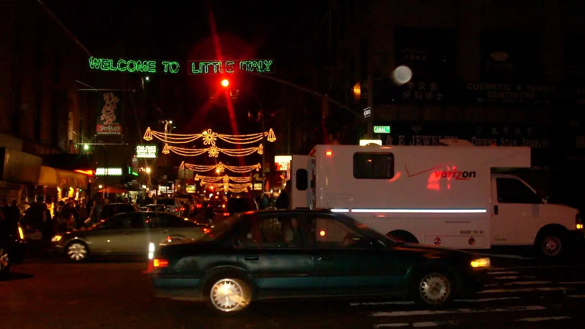 「WELCOME TO LITTLE ITALY」のネオンサインとイルミネーションが輝く夜のニューヨークの通り。乗用車とVerizonの白いサービスバンが停車している。
