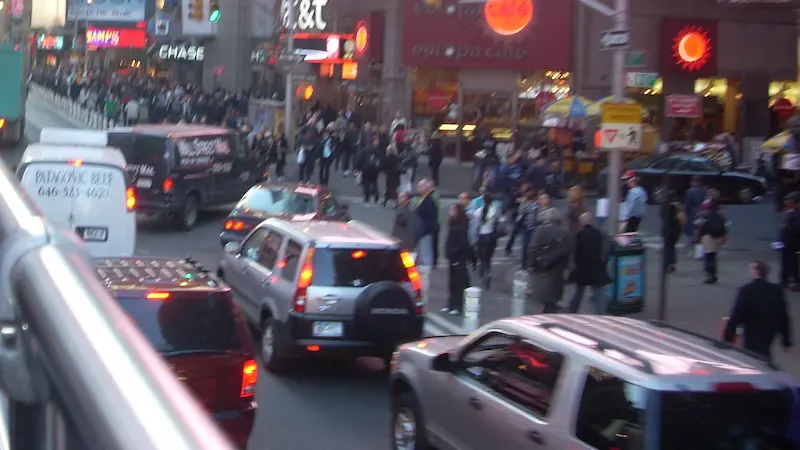 ニューヨークのタイムズ・スクエアと思われる賑やかな交差点で、車が行き交い、多くの人々が歩いている様子。