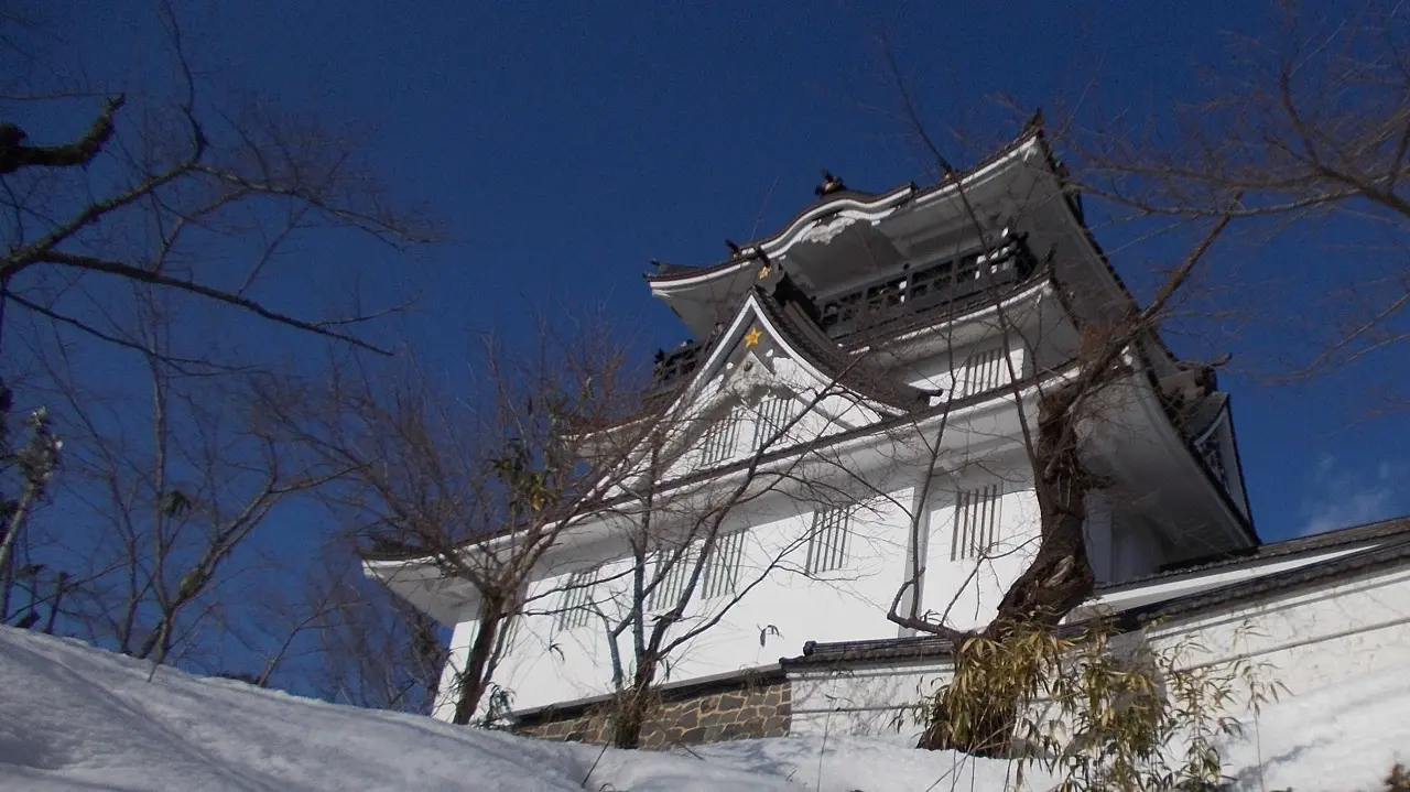 雪に包まれた日本の伝統的な城。白壁と黒瓦の屋根が青空に映え、葉を落とした木々が冬の静けさを際立たせている。一寸法師の物語の舞台を思わせる幻想的な風景。