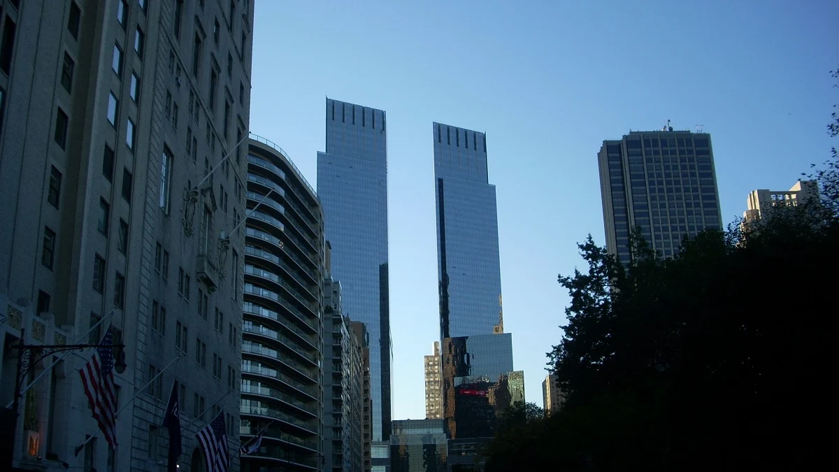 ニューヨーク、コロンバス・サークルの夕暮れ時の風景。中央には空を映し出すガラス張りのドイツ銀行センターのツインタワーがそびえ立ち、その左側には特徴的な曲線を持つ200 セントラル・パーク・サウスのビルが見えます。手前には街路樹のシルエットがあり、都会的で洗練されたマンハッタンの街並みを捉えています。